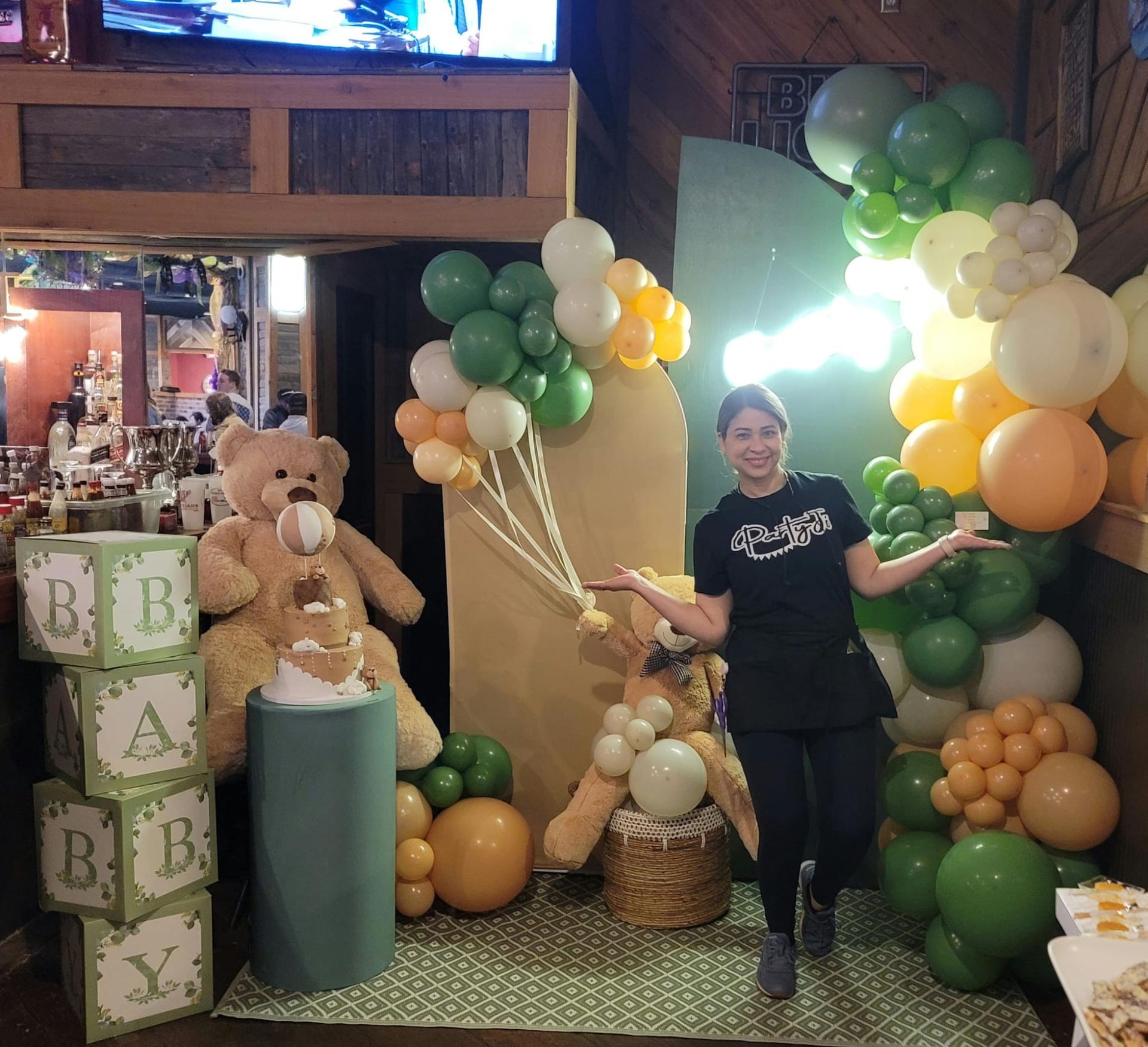 Woman posing at a baby shower with teddy bears and green, yellow, white balloons.