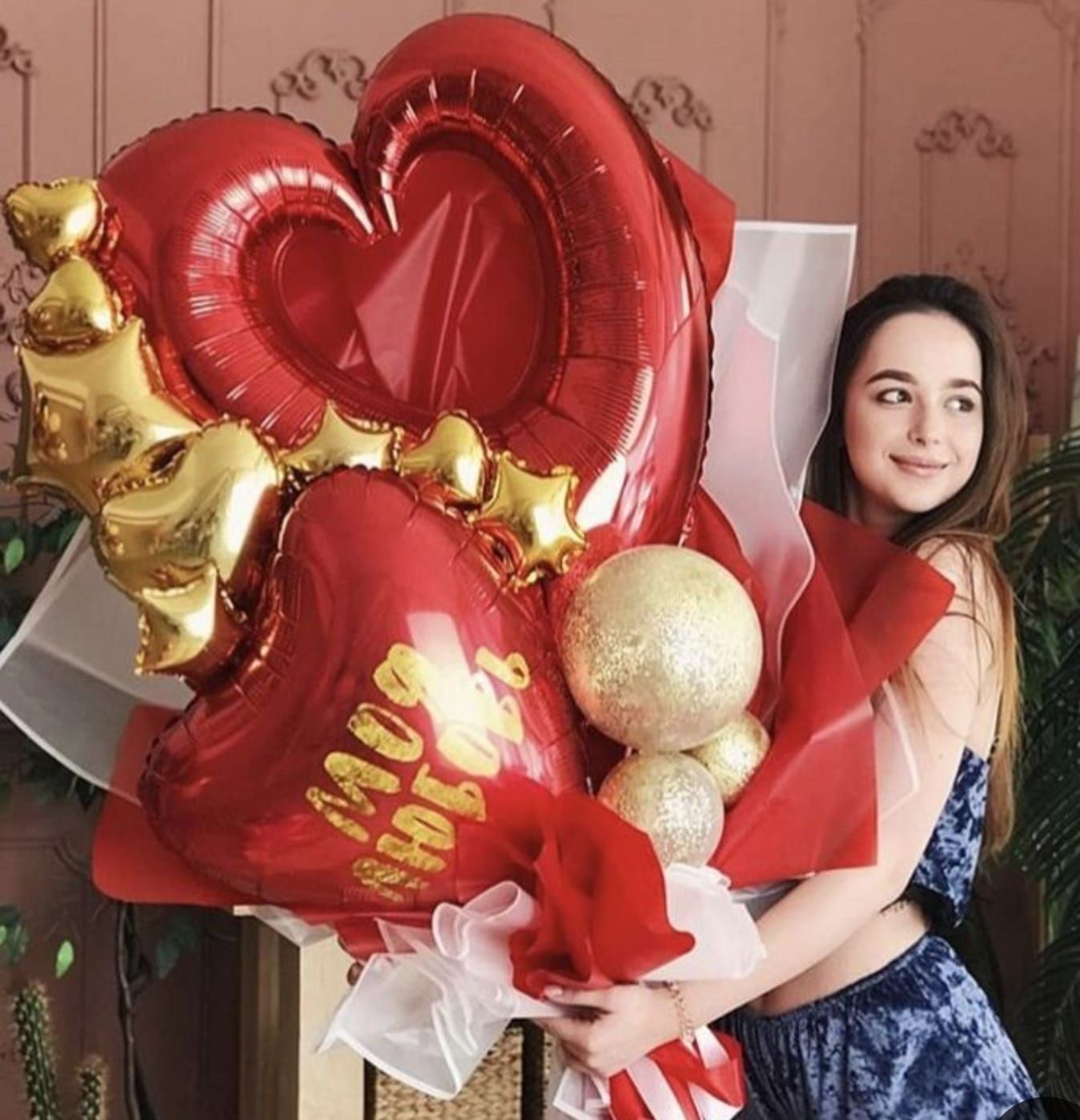 Woman holding a bouquet of red and gold heart-shaped balloons with star accents in a decorated room.